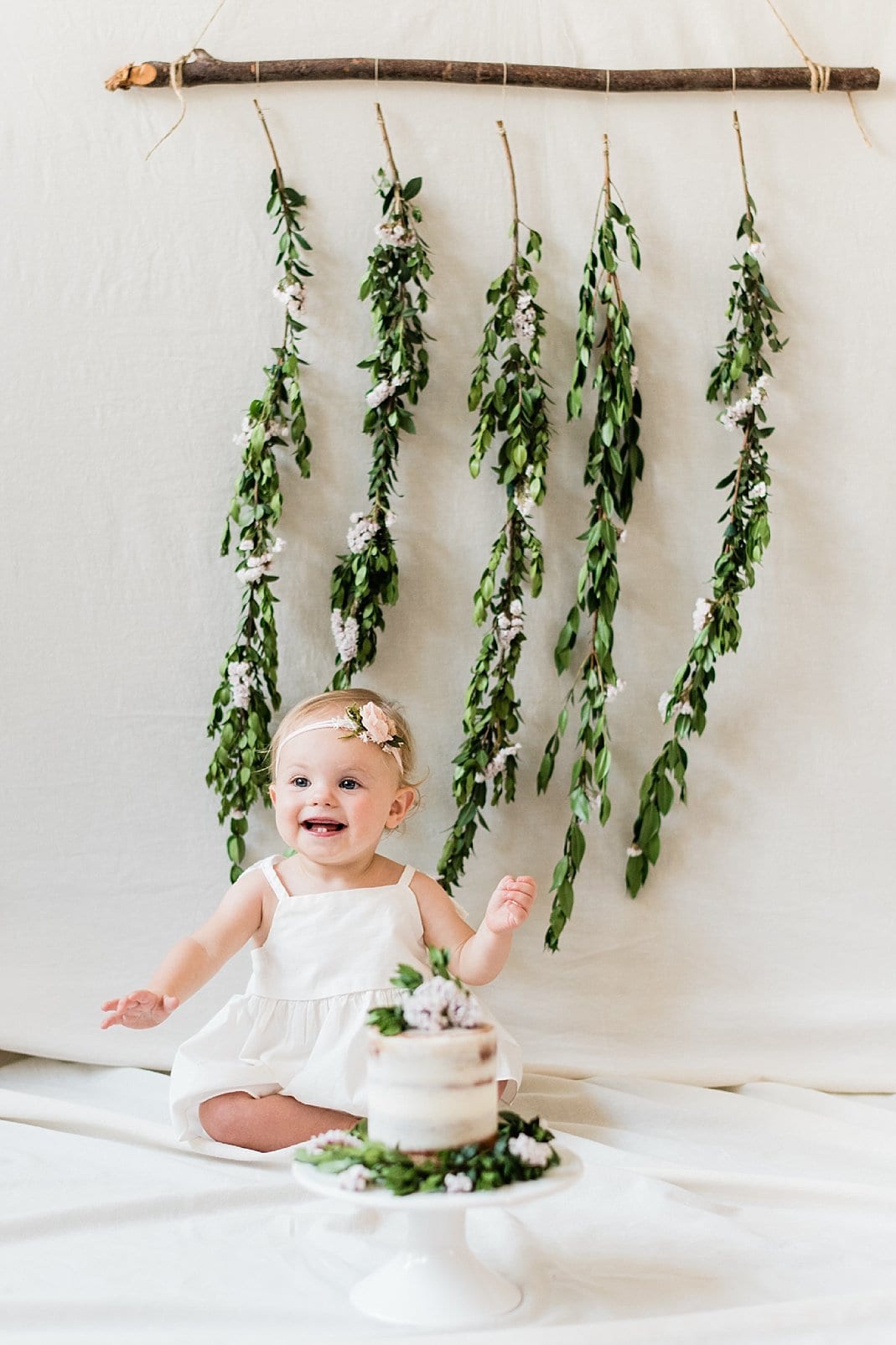 Raleigh cake smash for 1 year old girl in front of backdrop with greenery hanging down photo