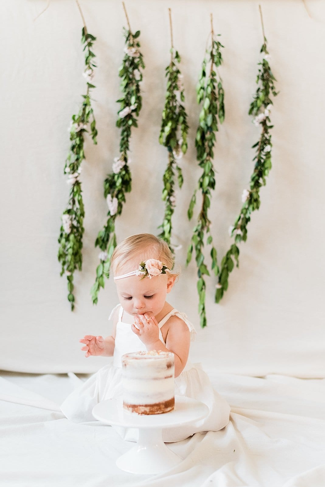 Wake Forest 1 year old doing an indoor cake smash in front of greenery backdrop photo