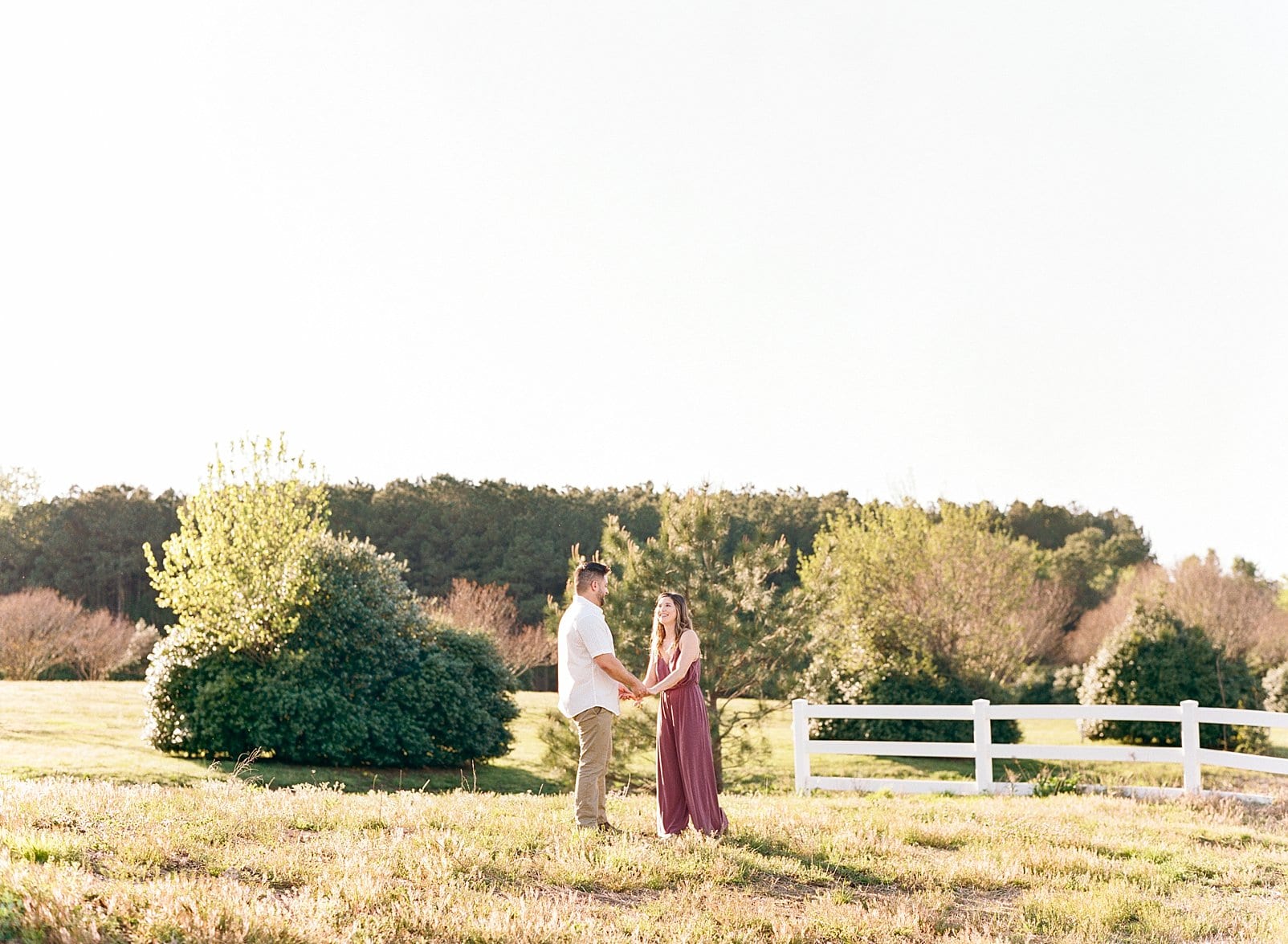 Raleigh, NC engagement shoot with couple holding hands in a field with a white fence behind them photo