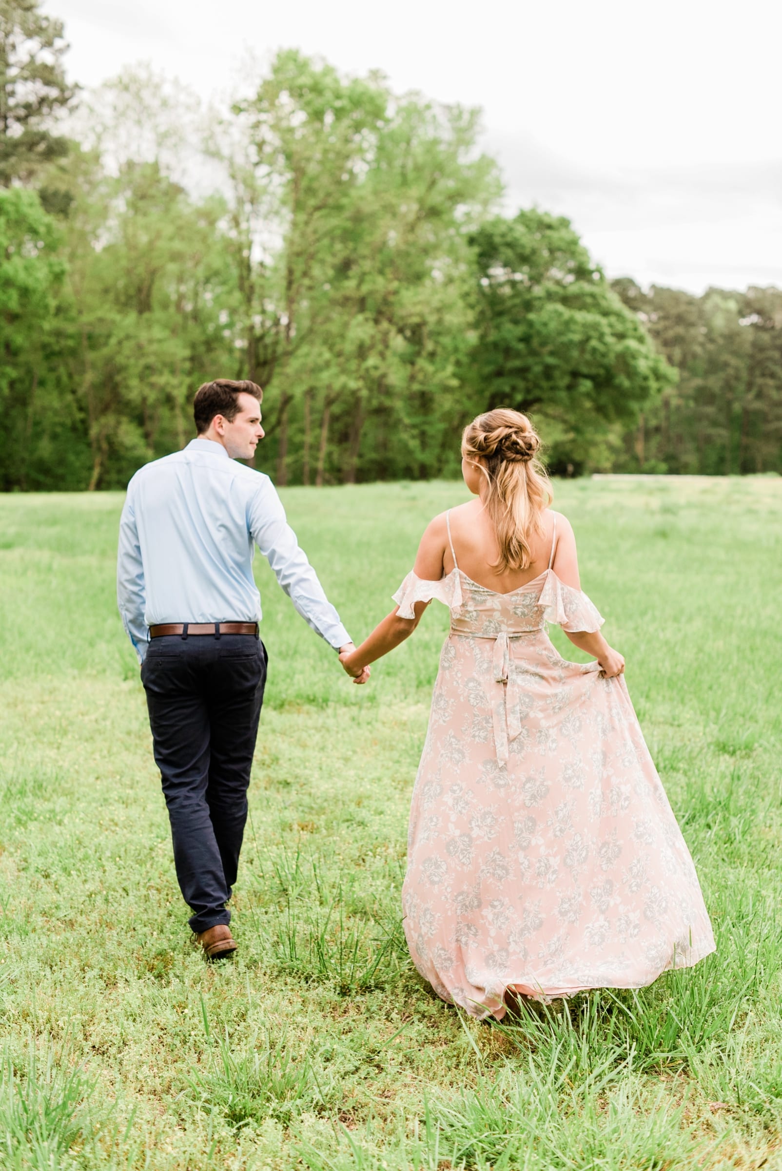 Engagement session with bride a long blush pink dress, holding it up while her groom leads her through a field photo