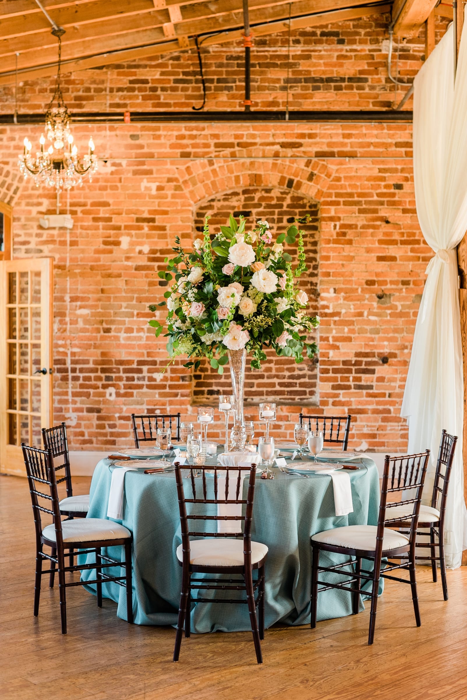 Melrose knitting mill reception with tall floral installation on a clear glass vase with a light blue table cloth and wooden chairs with white cushions photo