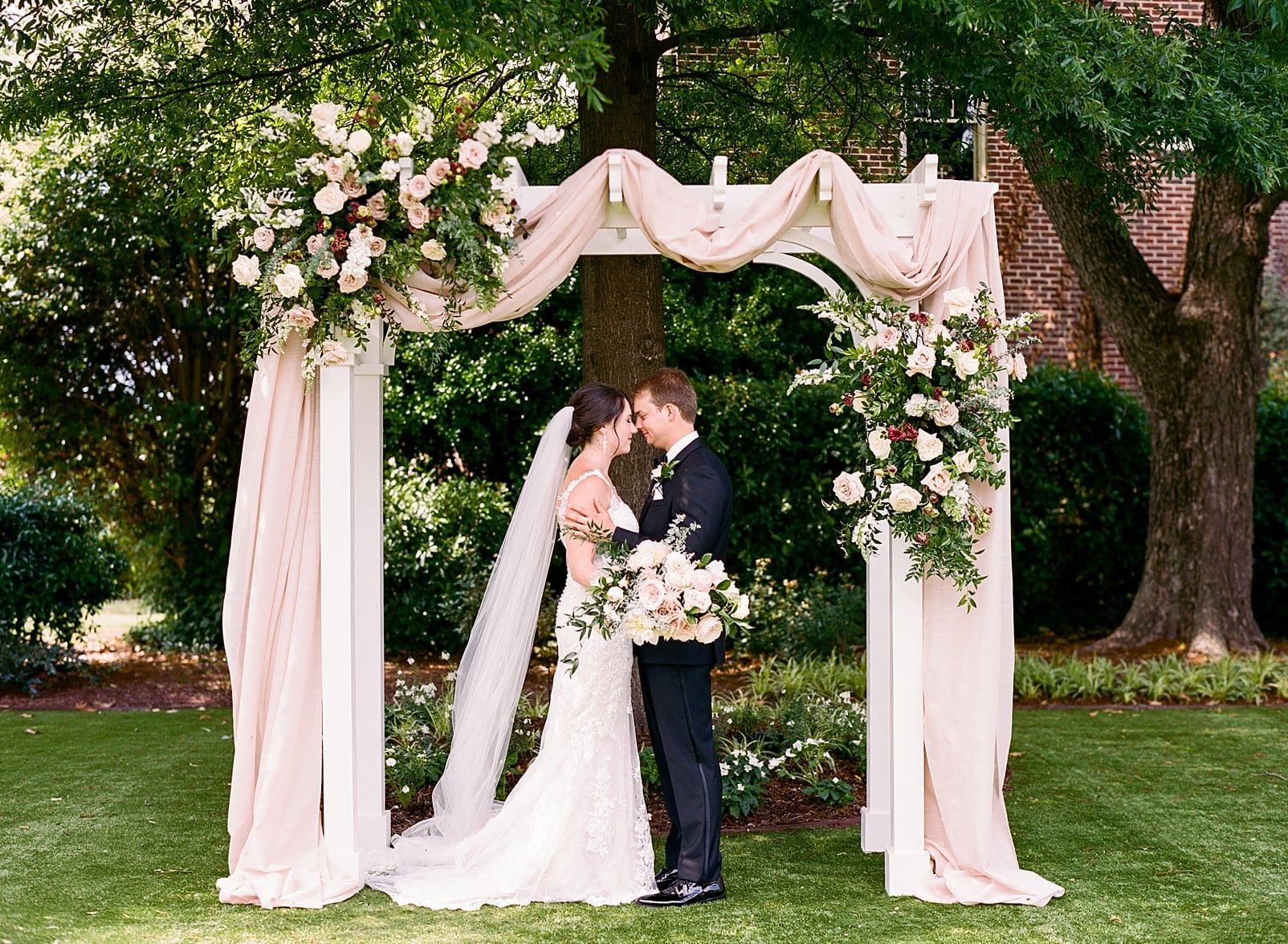 Merrimon Wynne bride and groom under the arbor at the ceremony sit. Arbor is draped with a long cloth and floral installments at the corners photo