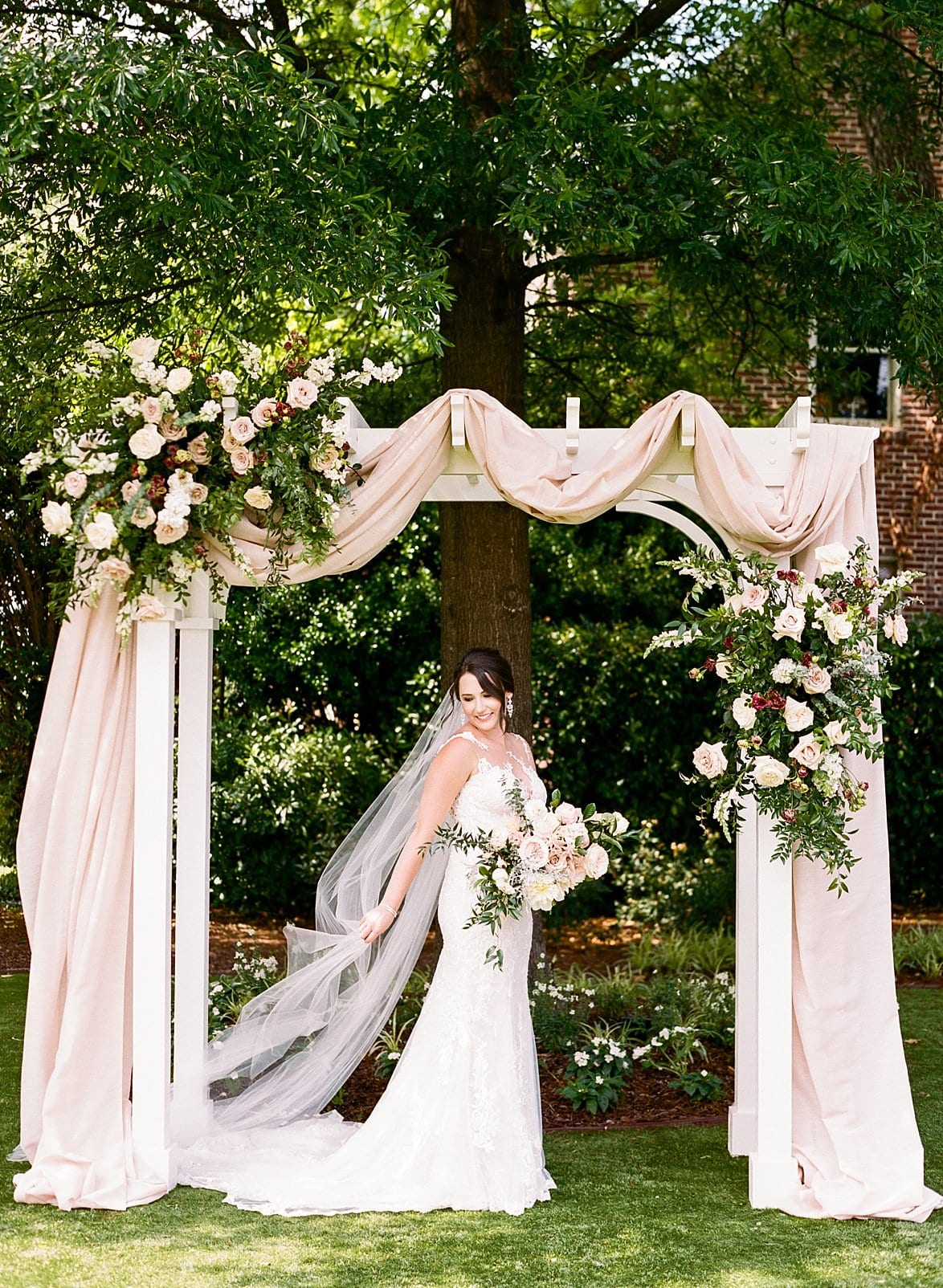 Merrimon Wynne bride standing under the white arbor with her veil and train behind her photo