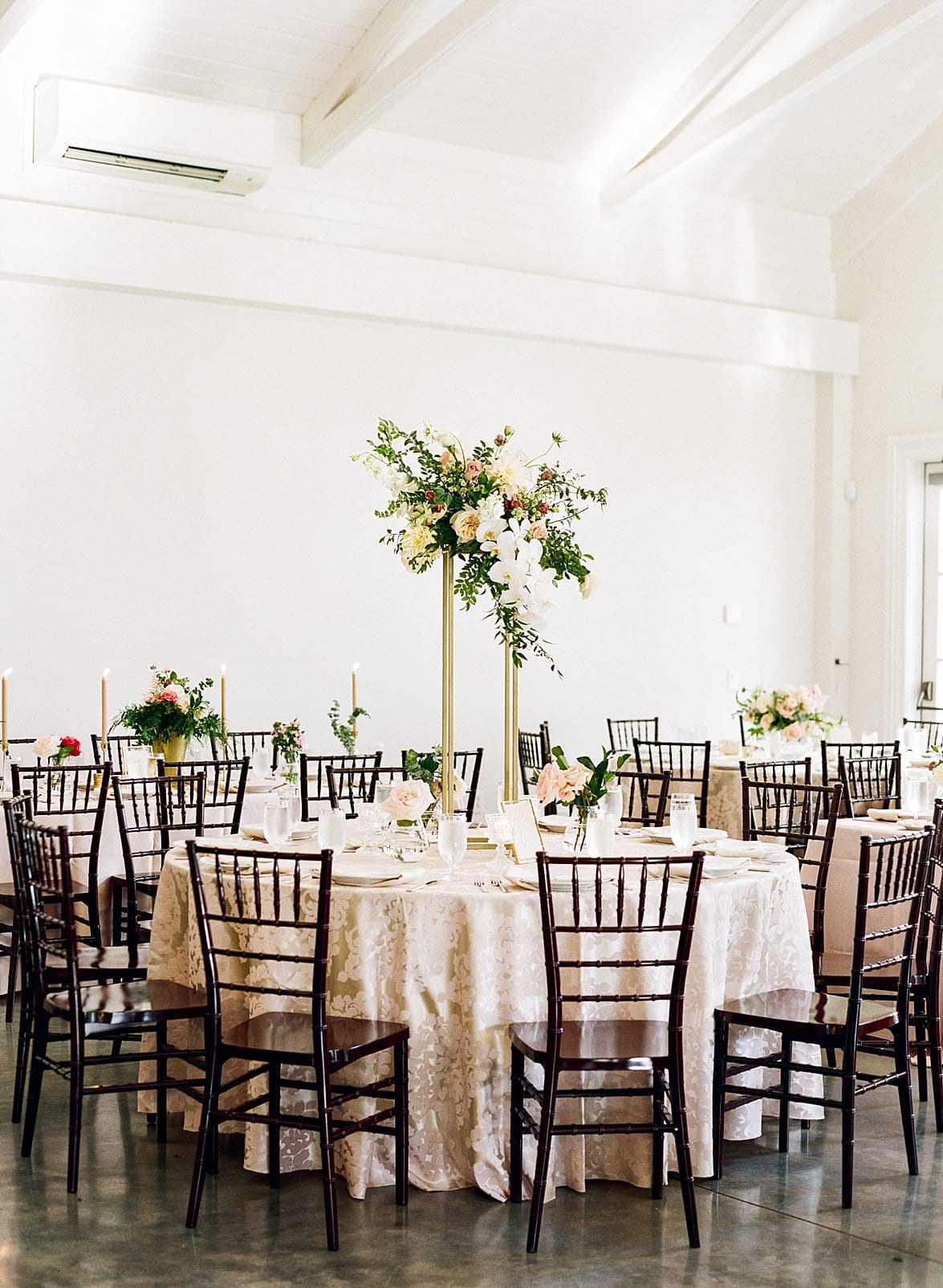 Merrimon Wynne indoor wedding reception with cream table clothes and brown wooden chairs and tall floral centerpieces on gold poles photo