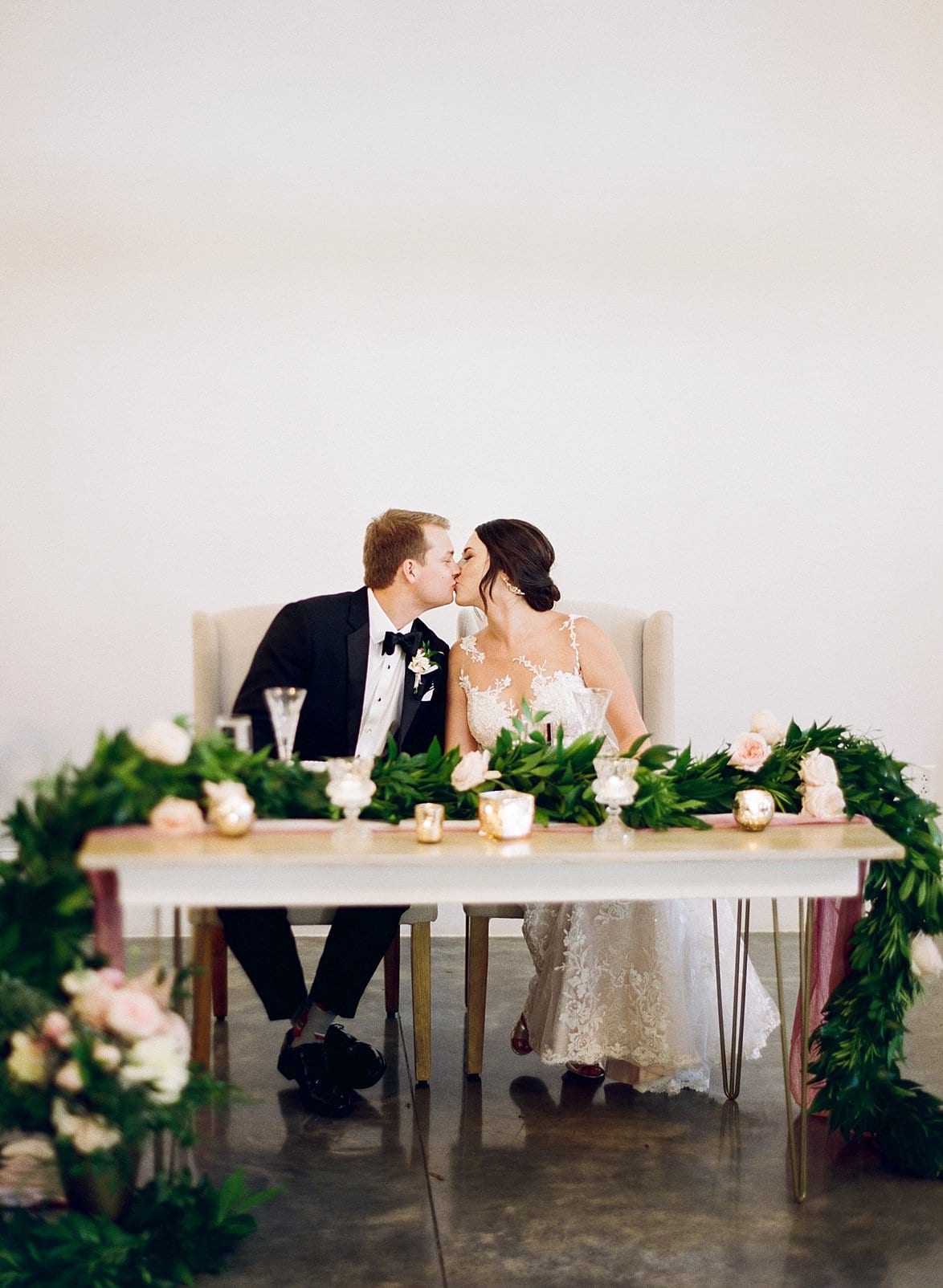 Merrimon Wynne bride and groom kissing while kissing at the sweetheart table draped with greenery and candles across it photo