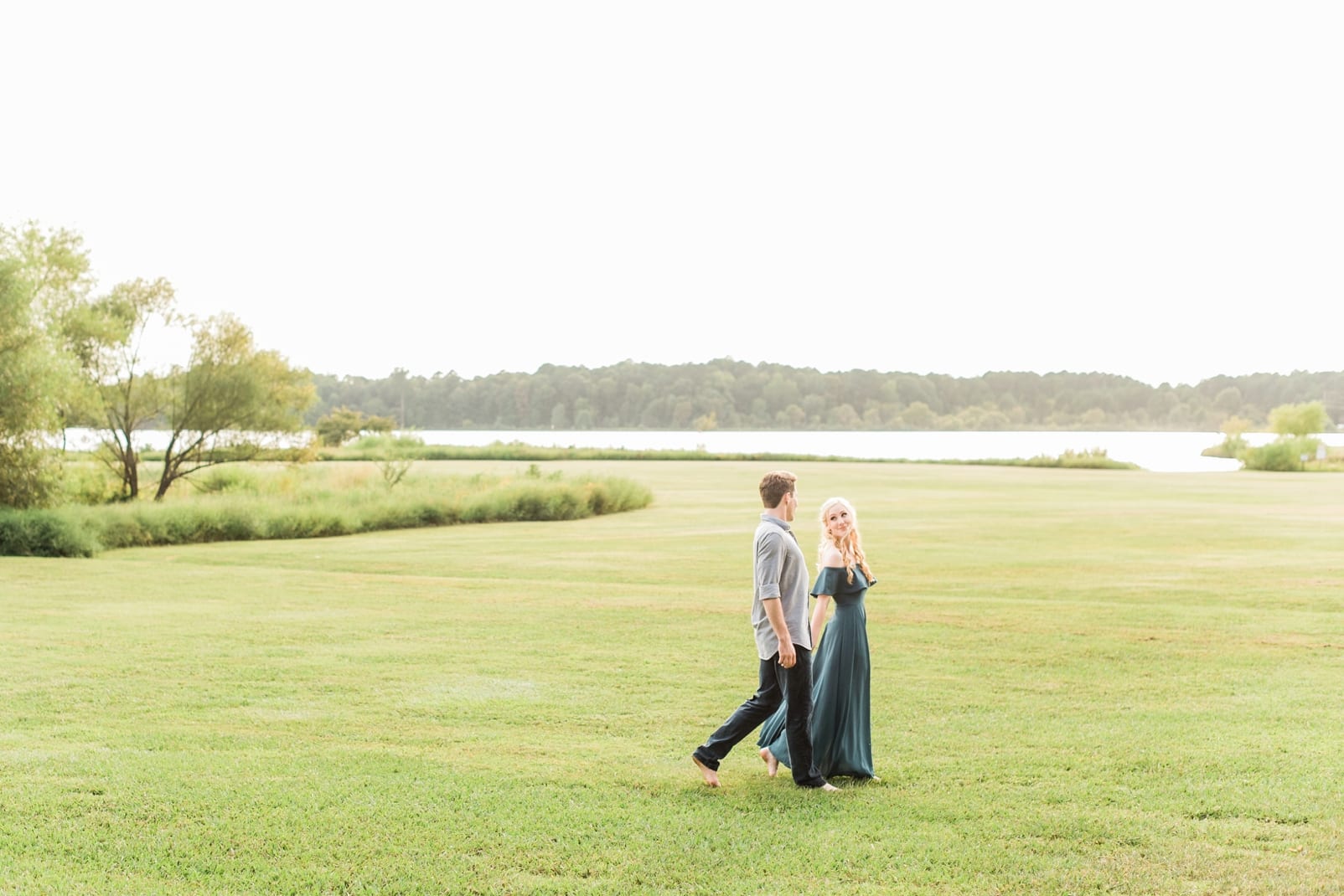 Raleigh couple walking through an open field with her in a long off the shoulder green dress and a lake in the background photo